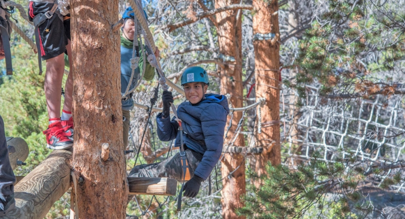 A student wearing safety gear is secured by ropes as they smile at the camera during a ropes course. 